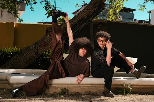 Two fashionable men with curly hair pose playfully outdoors on a sunny day.